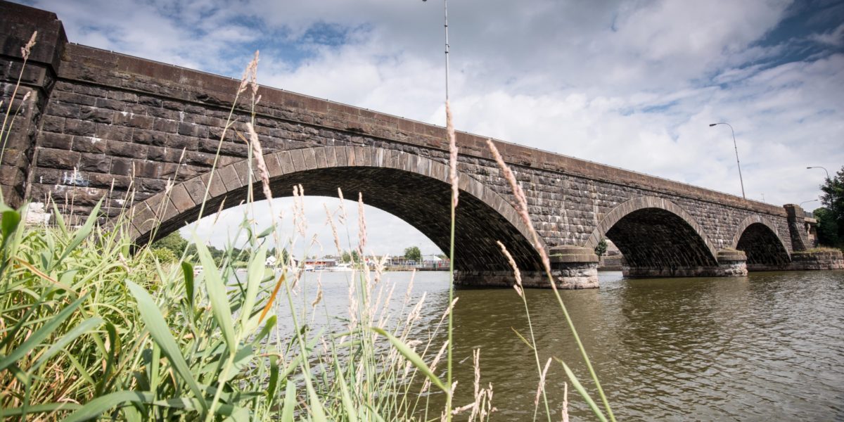 The historic stone bridge over the River Bann in Banbridge, Northern Ireland, with reflections on the calm water and lush trees lining the banks.